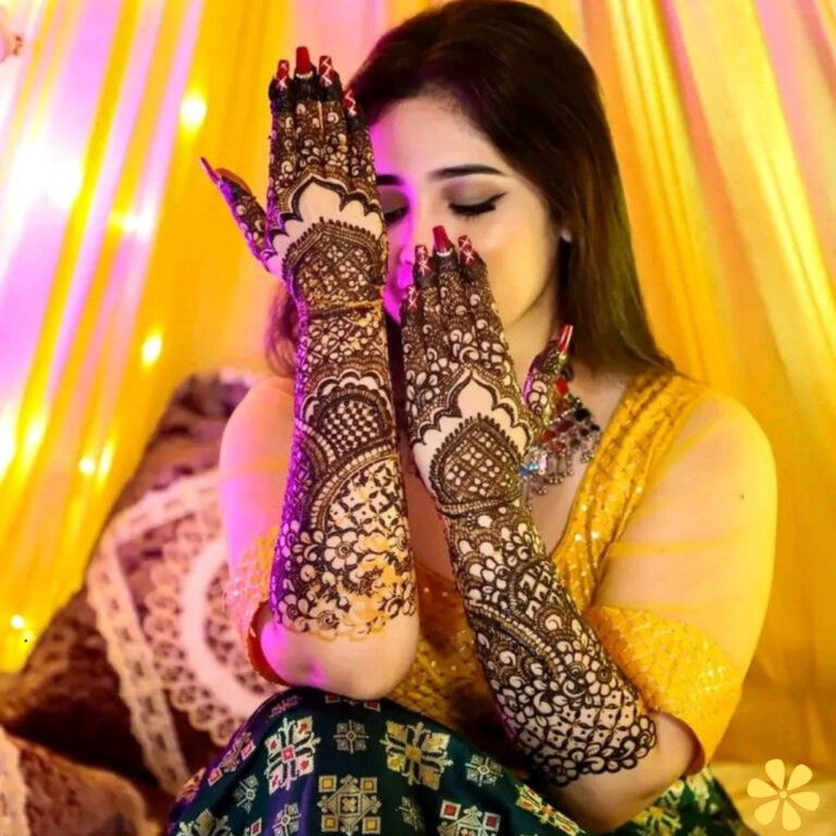 A woman in a yellow outfit with intricate henna on her arms poses with hands covering her face, surrounded by soft lights.