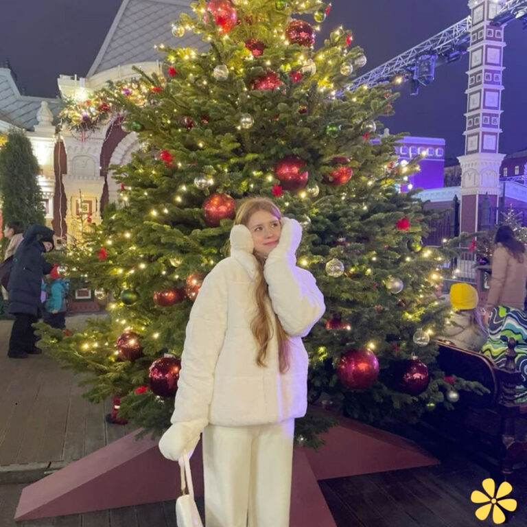 A young woman in a fluffy white coat stands by a festive Christmas tree adorned with lights and ornaments.