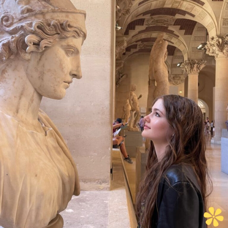 A young woman gazes up at a marble statue in a grand museum hall, her expression thoughtful and curious.