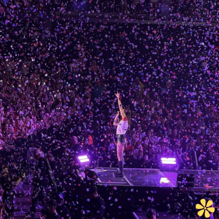 A performer stands on stage amidst a shower of colorful confetti, with fans cheering in the background.