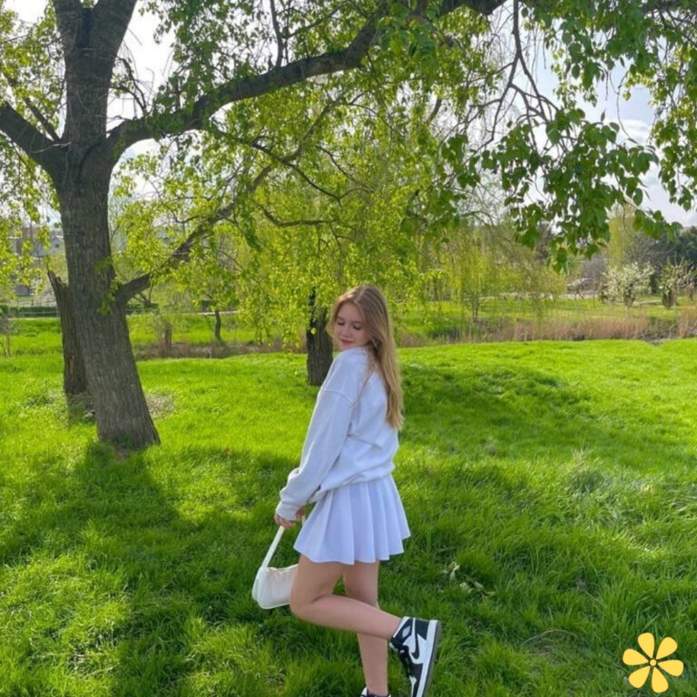 A young woman in a white sweater and skirt poses playfully in a sunny green park.