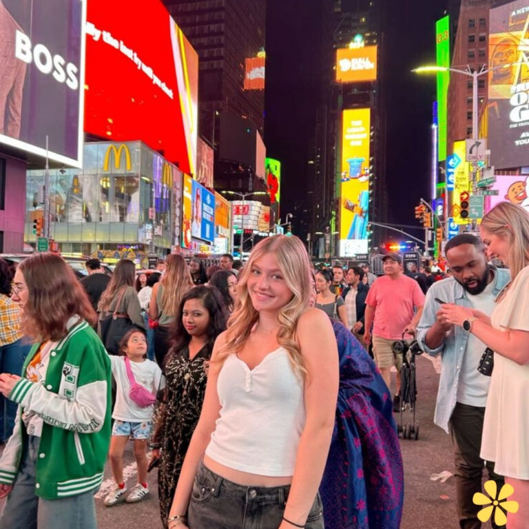 A young woman with blonde hair smiles in a crowded Times Square at night, illuminated by neon signs.