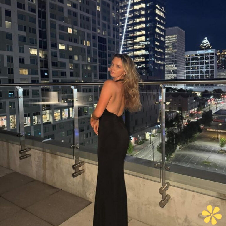 Woman in a sleek black dress poses confidently on a rooftop at night, city lights glimmering behind her.