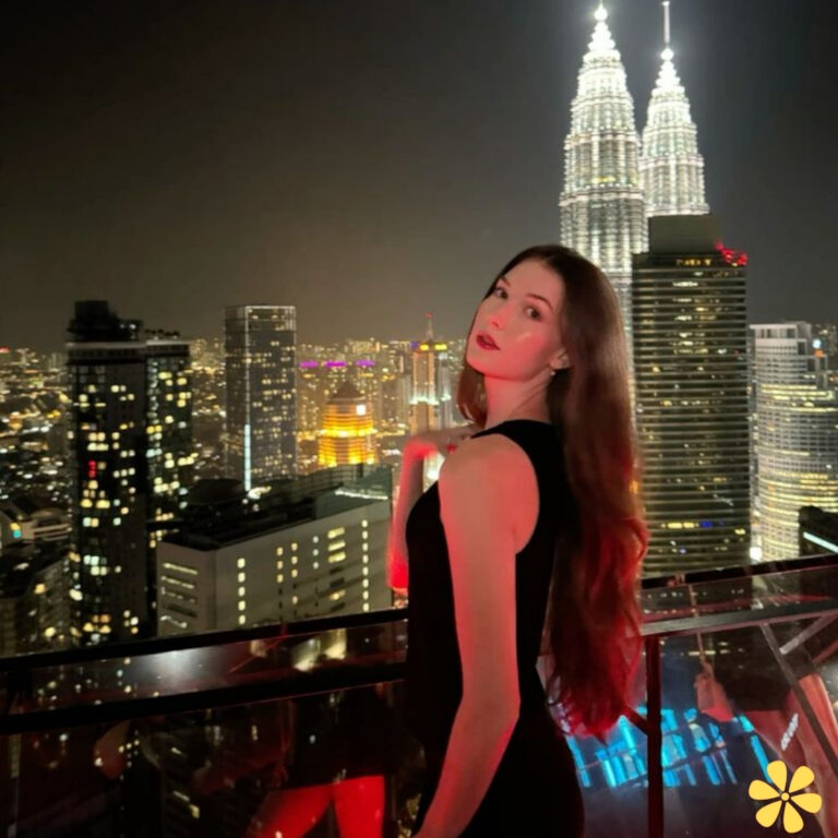 Woman with long hair poses elegantly against a city skyline at night, featuring bright lights and iconic towers.