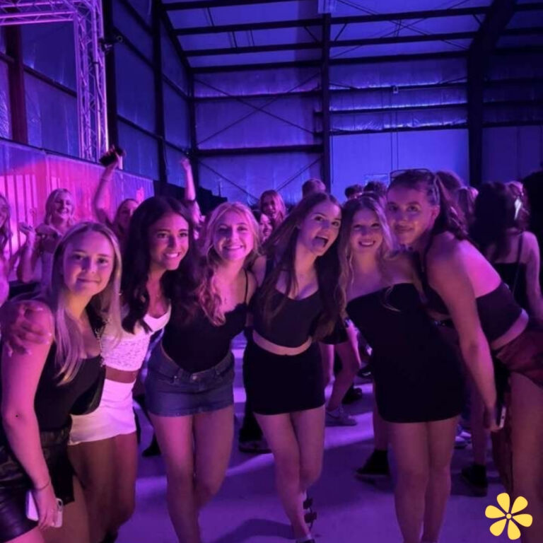 Six friends smile and pose together at a lively event, vibrant lights shining behind them.