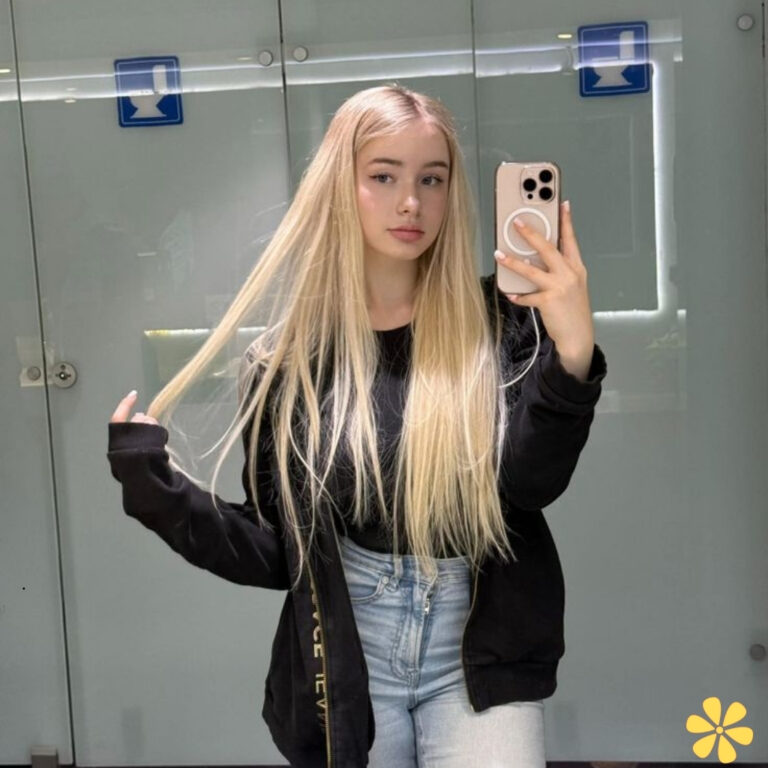 Young woman with long blonde hair taking a selfie in a mirror, wearing a black jacket and jeans.