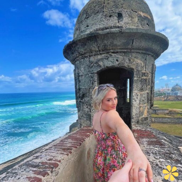 Woman in floral dress stands on a fort, holding hands with someone, ocean waves behind her.