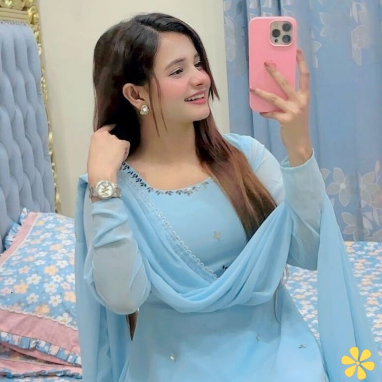 Woman in a light blue outfit smiles while taking a selfie, hair down, in a cozy room.