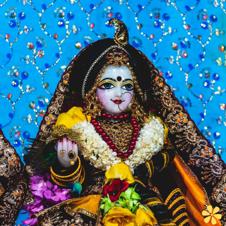 A beautifully adorned idol with intricate jewelry and colorful flowers against a vibrant blue backdrop.
