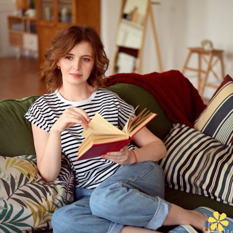 Woman with curly hair reading a book on a cozy couch, surrounded by cushions and a warm setting.