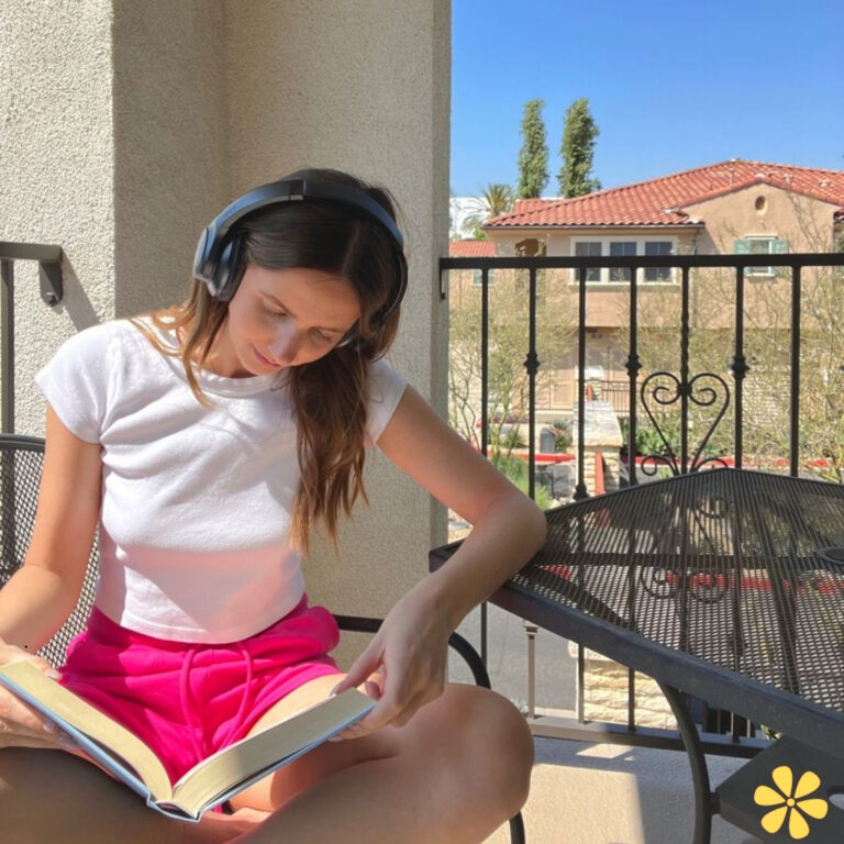 Young woman in headphones reading a book on a balcony in bright sunlight.