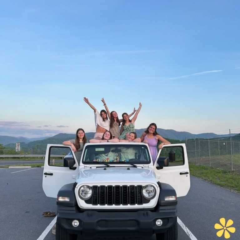 Group of friends joyfully posing in a Jeep, mountains in the background, sunny day.