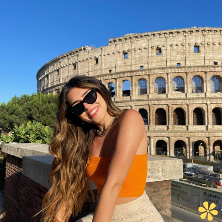 Woman in orange top smiles in front of the Colosseum, sunny day, sunglasses on, long hair flowing.