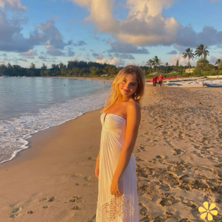 A woman stands on a sandy beach at sunset, wearing a flowing white dress, with soft waves lapping at her feet.