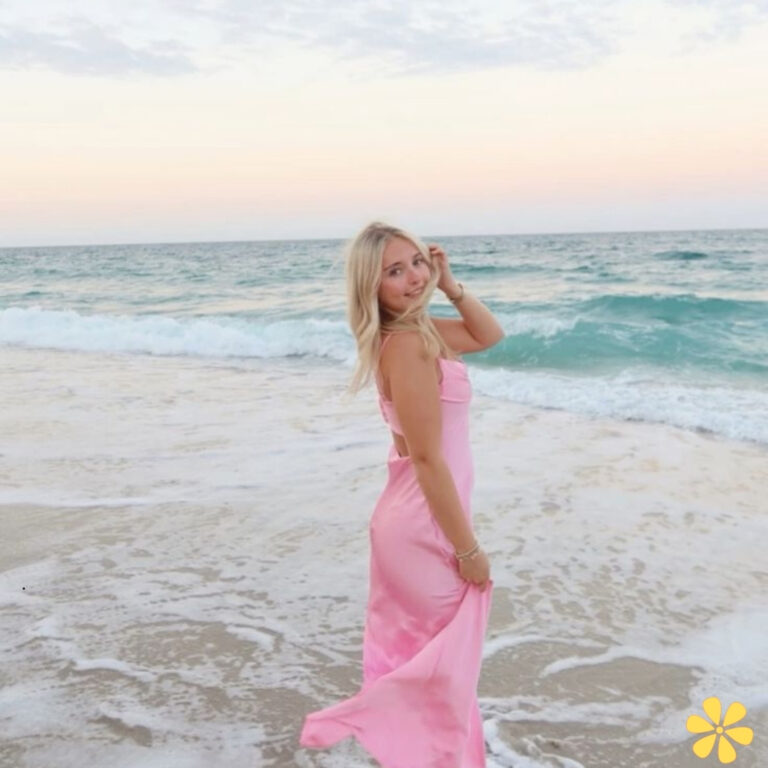 Woman in a flowing pink dress, playfully touching her hair at the beach during sunset.