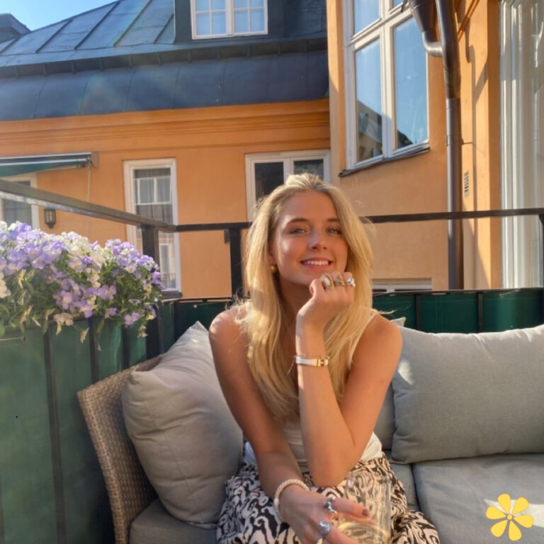 Young woman sitting on a couch with a smile, wearing a tank top and patterned pants, enjoying a sunny day.