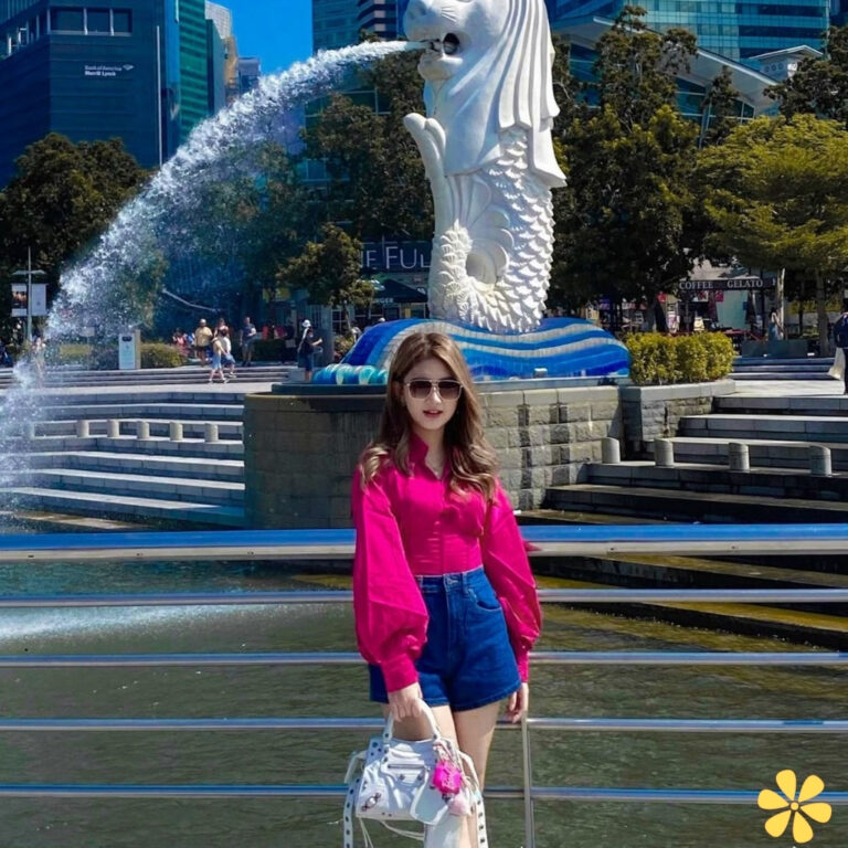 Woman in pink top and shorts poses by fountain with lion statue, sun shining.