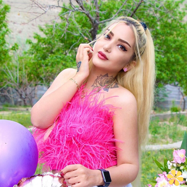 A woman poses confidently in a vibrant pink feather outfit, surrounded by greenery and balloons.