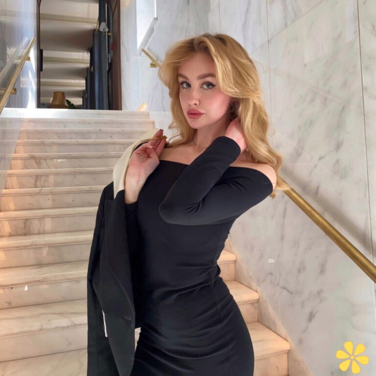 A woman poses confidently on marble stairs, wearing a sleek black off-shoulder dress and holding a blazer.