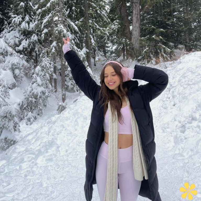 Woman in winter attire with arms raised, smiling in snowy forest.