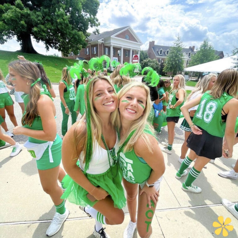Two smiling women dressed in green outfits with matching accessories, surrounded by a lively crowd.