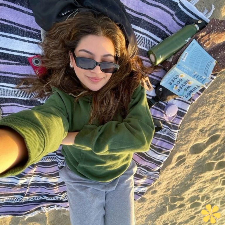 Woman lounging on a beach blanket, wearing sunglasses and a green sweater, smiling at the camera.