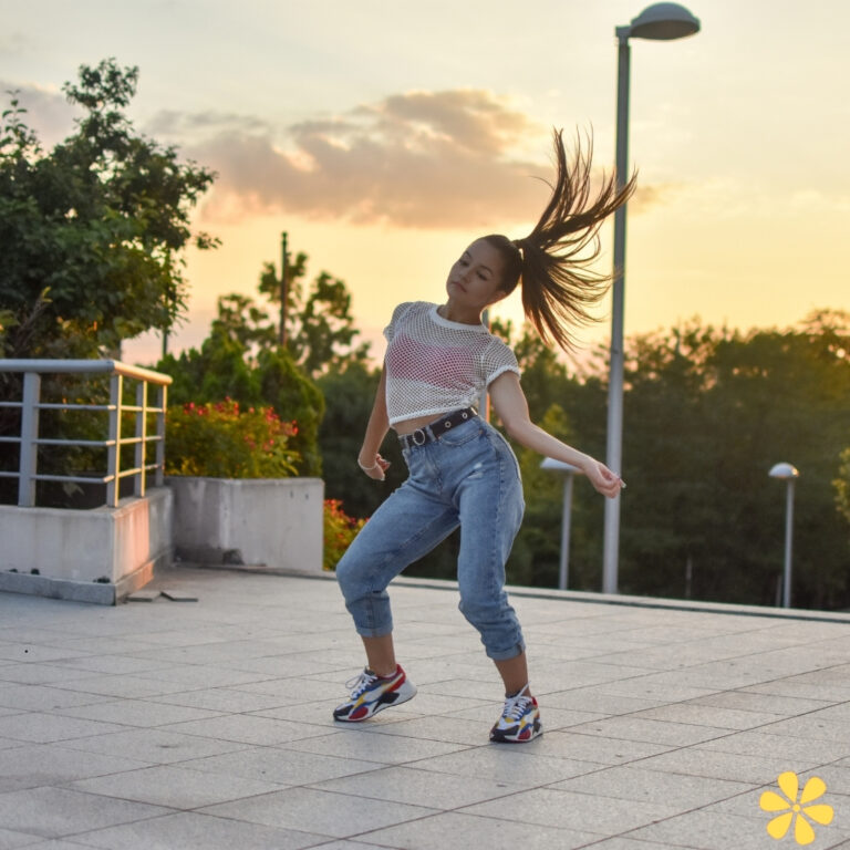 Young woman dancing outdoors at sunset, hair flying, wearing a mesh top and high-waisted jeans.
