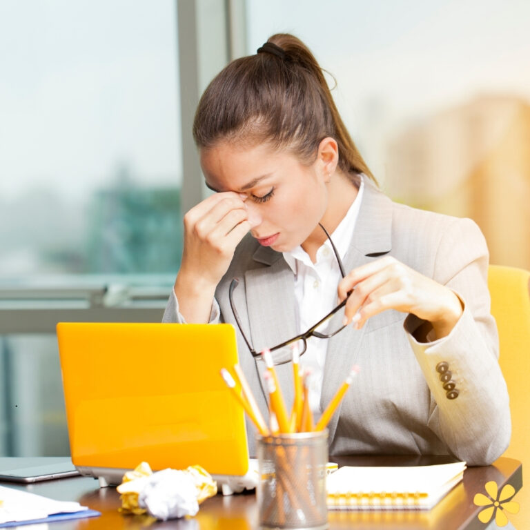 Frustrated woman in a suit holds glasses, resting forehead on hand at her desk with a laptop.