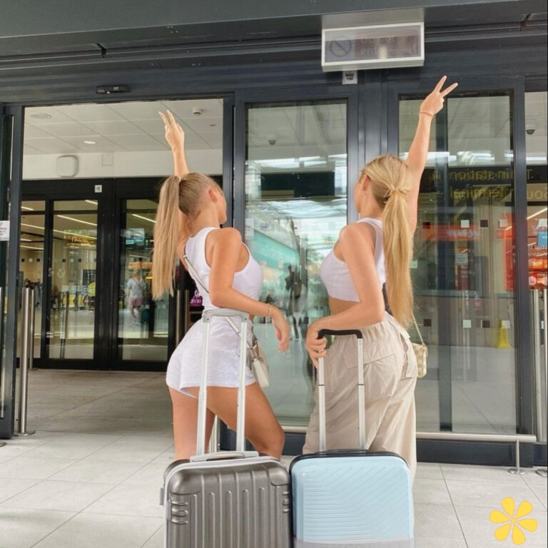 Two friends pose with luggage at an airport, giving peace signs.