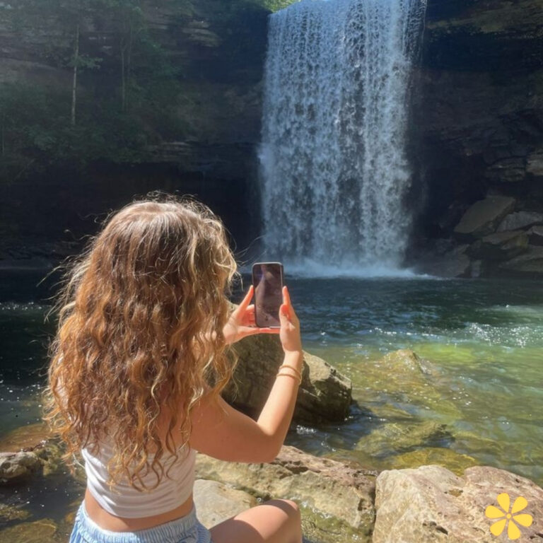 A person with curly hair takes a photo of a waterfall while sitting on rocks by a serene pool.