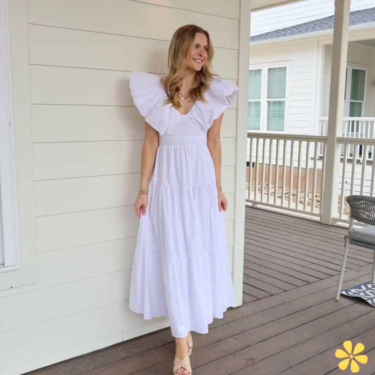 Woman in a white dress with ruffled sleeves, standing on a porch, smiling.