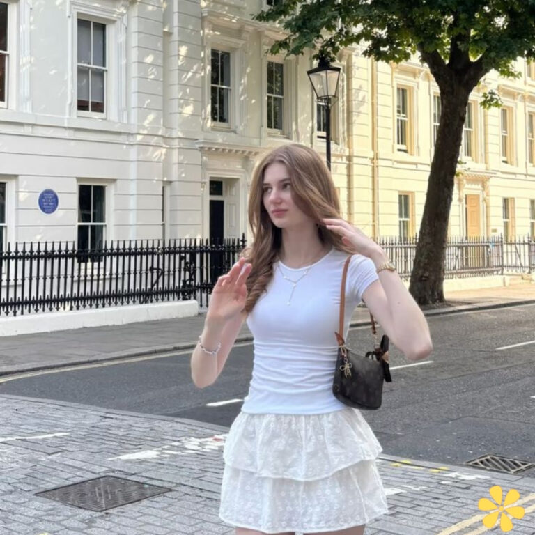 Young woman waves while walking on a street with white buildings in the background.