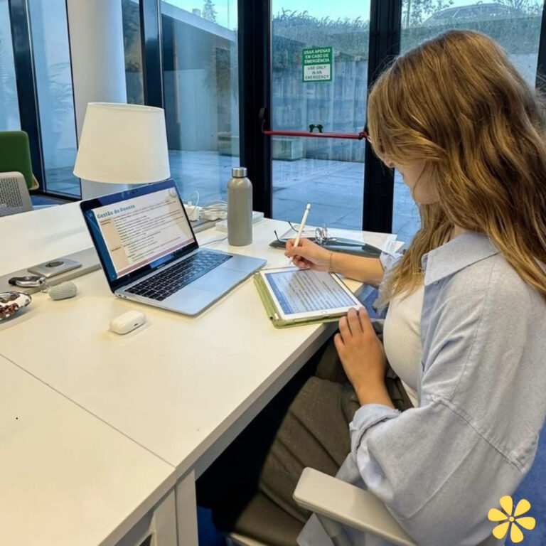 Woman writing notes beside laptop in bright workspace with greenery outside.