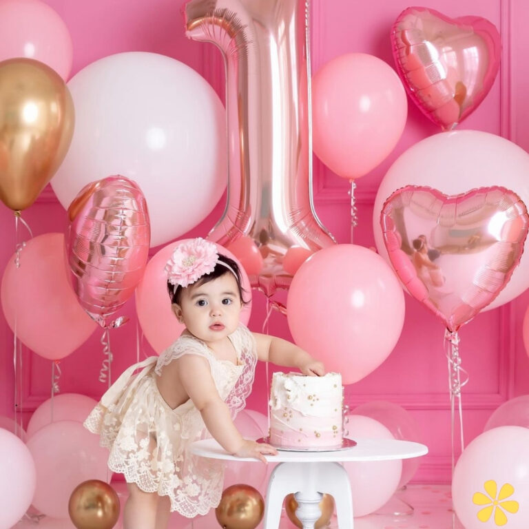 A cute baby girl in a lace dress plays with a small cake surrounded by pink and gold balloons.