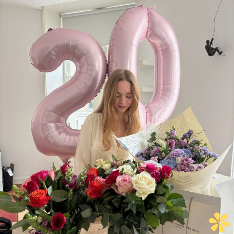 Young woman with long hair joyfully arranging flowers, surrounded by large pink '20' balloons.