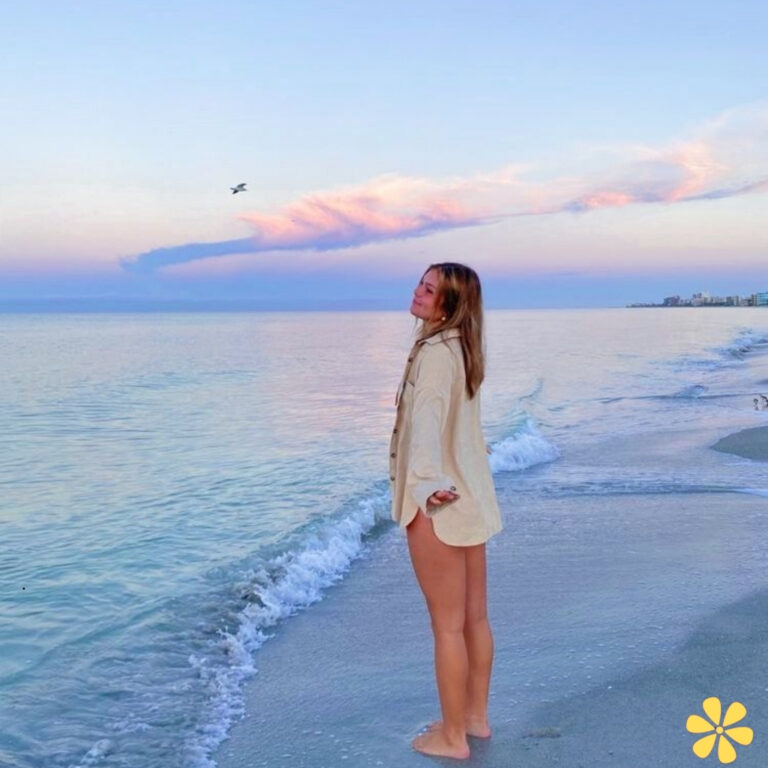 A woman stands barefoot by the shore, feeling the gentle waves lap at her feet, with pastel skies above.
