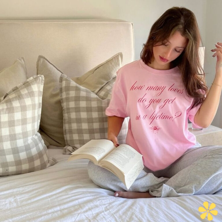 Young woman in a pink shirt, sitting on a bed with pillows, reading a book.