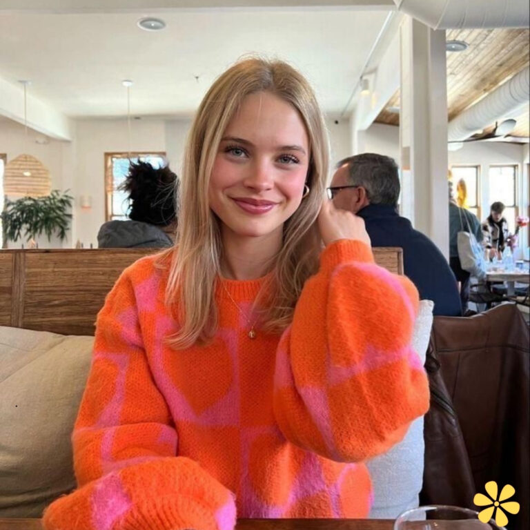 Smiling woman in vibrant orange and pink sweater sits at a cozy café table.