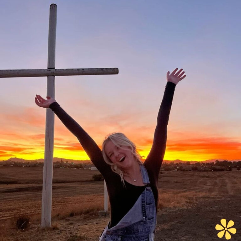 A woman with arms raised joyfully, standing in front of a cross against a vibrant sunset.