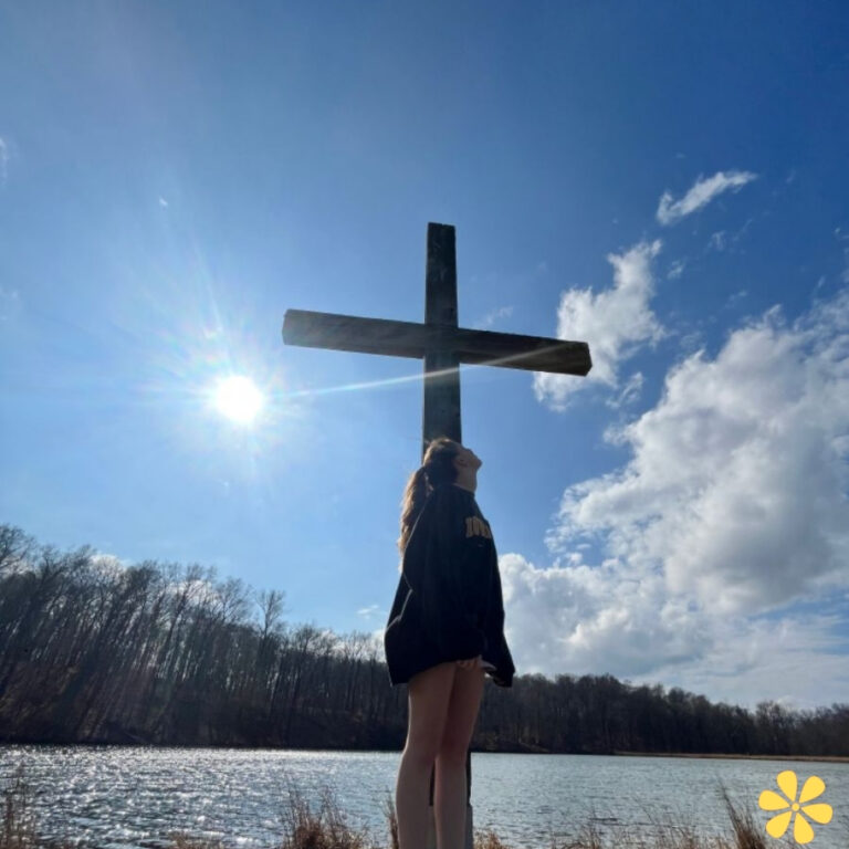 A woman stands by a large cross near a serene lake, sunlight streaming down.