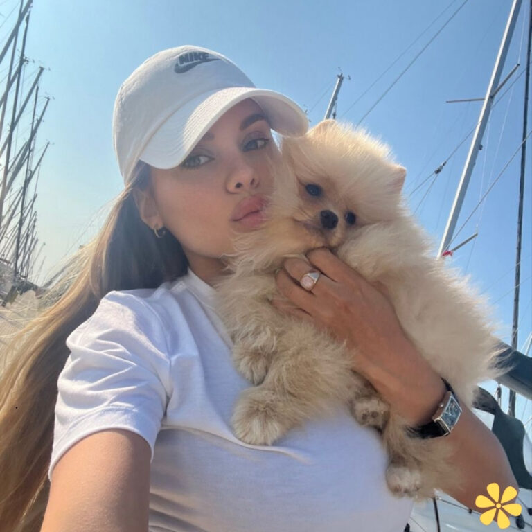 A woman in a white cap holds a fluffy pomeranian near a marina with sailboats.