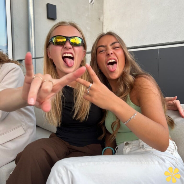 Two friends making funny faces and posing with peace signs, one wearing sunglasses.