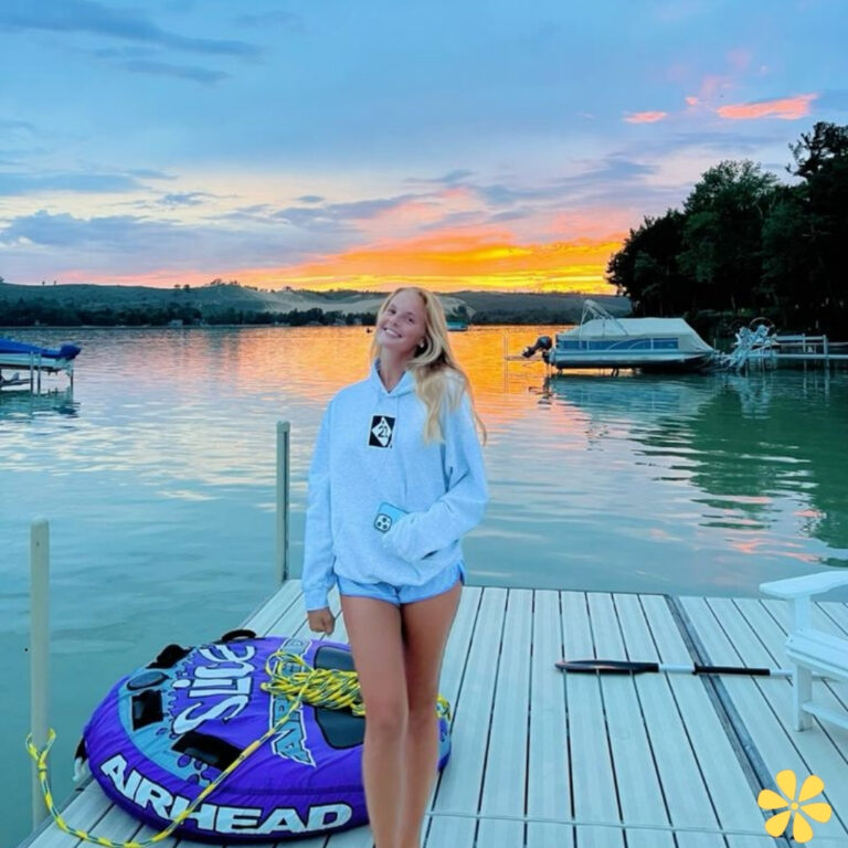 A woman in a hoodie stands on a dock, smiling at sunset over a lake.