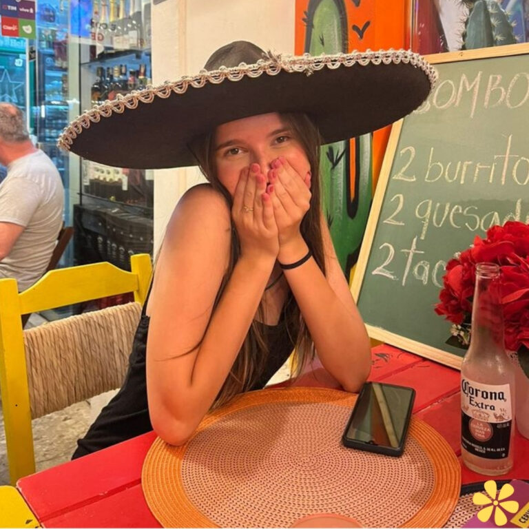 Young woman in a sombrero playfully covering her face, sitting at a colorful restaurant table with drinks and food.