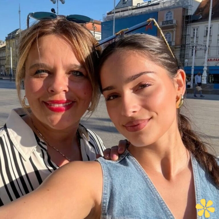 Two women smile together in a sunny plaza, one in a striped blouse, the other in a denim top.