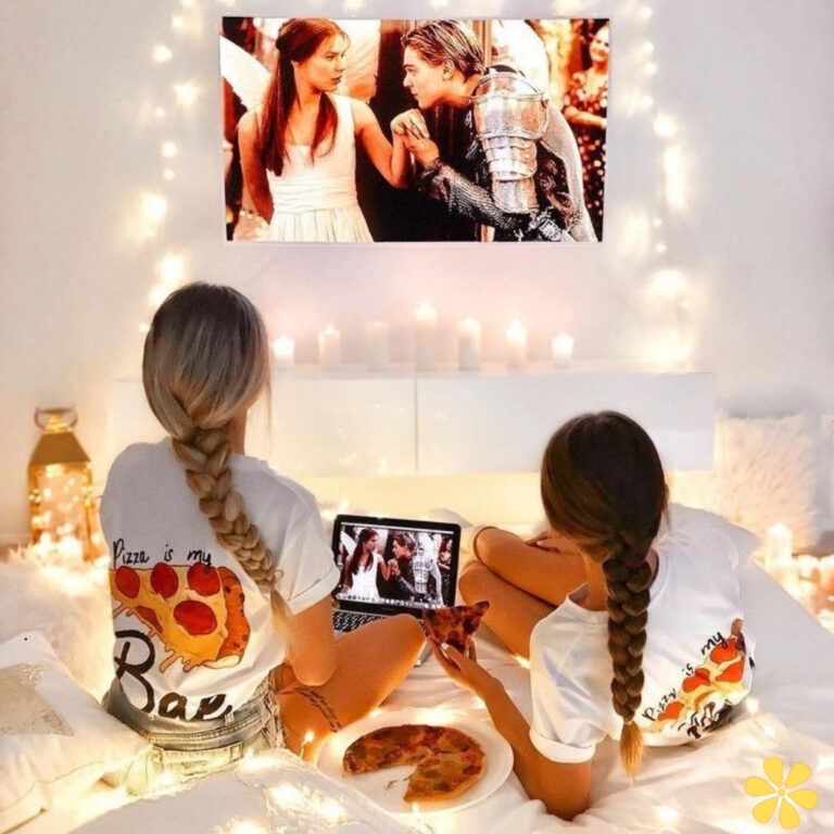 Two friends watch a romantic movie in cozy shirts while enjoying pizza on a bed with fairy lights.
