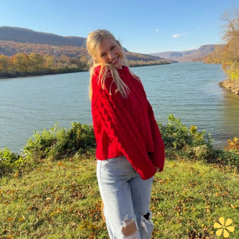 Smiling woman in a cozy red sweater stands by a tranquil river, mountains in the distance.