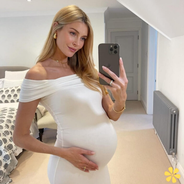 Pregnant woman in a white dress smiling while taking a mirror selfie.
