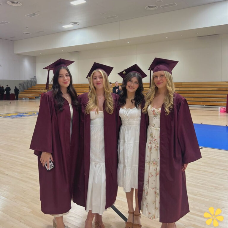 Four graduates in maroon gowns smile together in a gymnasium.