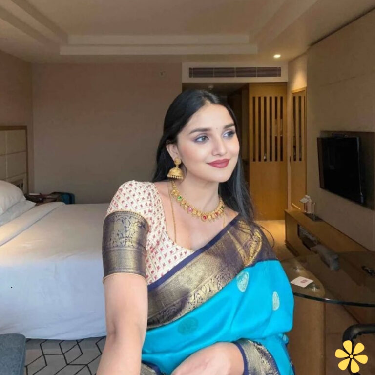 Woman in elegant blue saree and traditional jewelry, smiling softly in a cozy hotel room.